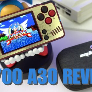 La Miyoo A30, una consola portátil de estilo retro, ocupa un lugar destacado. Su diseño dorado y negro muestra una pantalla en la que aparece Sonic the Hedgehog, rodeado de controles de juego clásicos que incluyen un D-pad, botones circulares y un stick analógico. El dispositivo está sostenido por una juguetona figurita con grandes dientes a la vista. En primer plano, una funda protectora negra con el logotipo de Miyoo A30. La imagen muestra el tamaño compacto y el atractivo nostálgico de la consola, destacando su portabilidad y sus capacidades de juego clásico. Un texto en negrita azul en la parte inferior reza &quot;MIYOO A30 REVIEW&quot;, indicando el propósito de la imagen. La composición general destaca la combinación de tecnología moderna y estética retro, lo que atrae a los aficionados a los videojuegos clásicos.