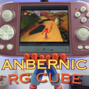 The image shows a handheld gaming console labeled "ANBERNIC RG CUBE" in the foreground. The console has a pinkish color and features a D-pad, two analog sticks, and several buttons, including X, Y, A, and B buttons. The screen of the console displays a gameplay scene from "Crash Bandicoot," where the character is running along a path collecting items. The console appears to be held up by a figure, possibly a toy or model, visible below the console. The background features a clean, modern setup with a blue-lit environment, and some other tech gadgets are partially visible.