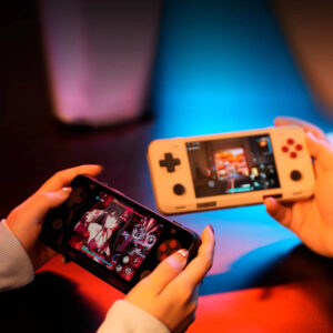 Two users holding black and retro beige AYANEO Pocket ACE consoles, each playing a different game under colorful ambient lighting in a cozy indoor setting.