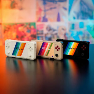 Three AYANEO Pocket ACE consoles—white, retro beige, and black—standing upright on a reflective black table, backed by a vivid and playful multicolored wall.