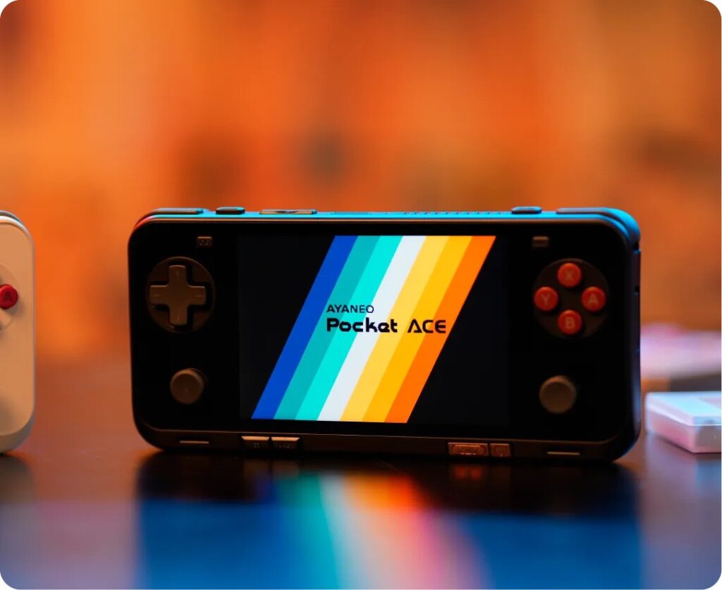 AYANEO Pocket ACE 1 A handheld gaming console, the AYANEO Pocket ACE, displayed on a table with a colorful gradient logo on its screen. The device features a D-pad, analog sticks, and action buttons, all illuminated in a warm ambient setting with a blurred orange background.