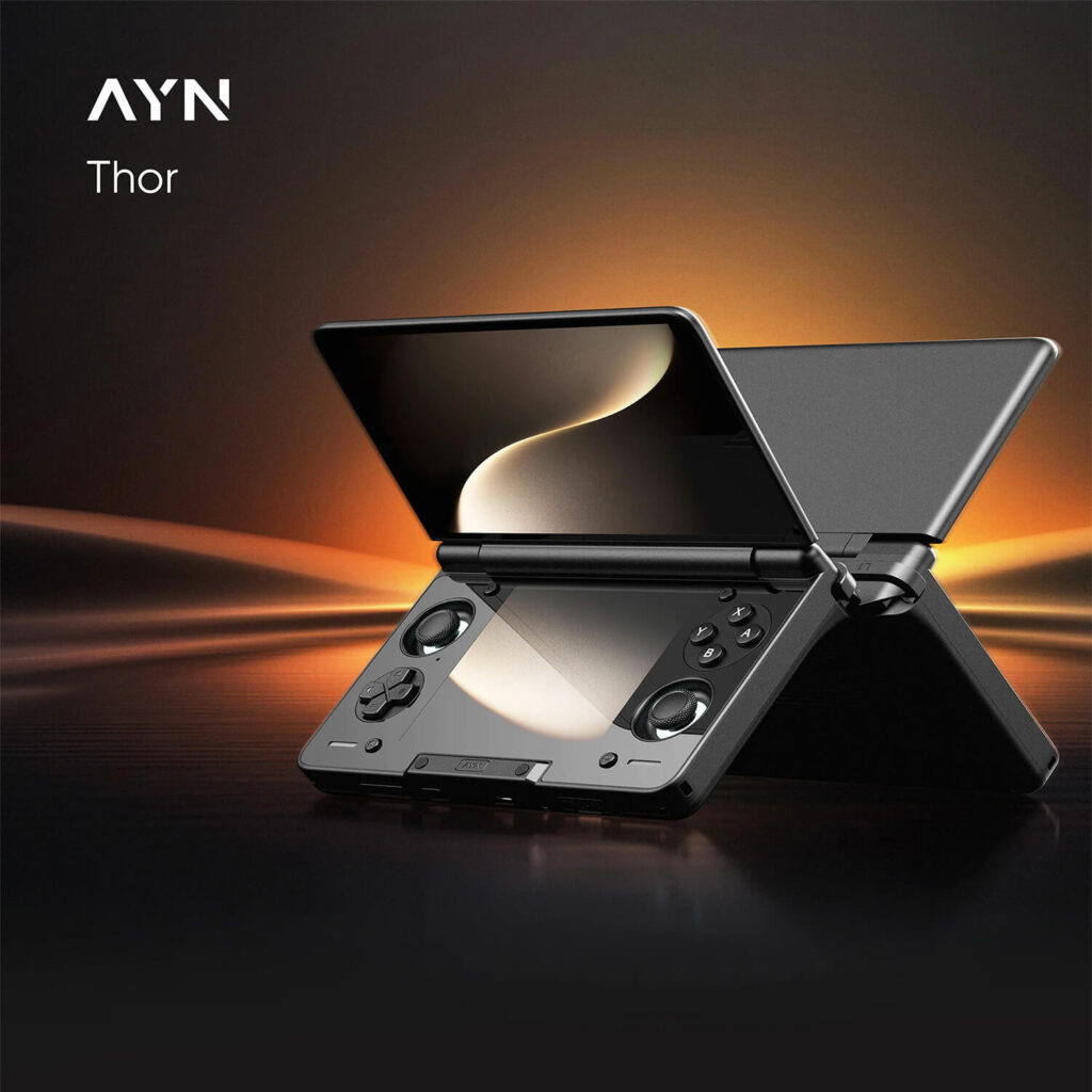 AYN Thor Lite, Base, Pro & MAX 1 A sleek, black AYN Thor handheld gaming console is shown angled upwards, with its clamshell design open like a book, against a dark, glowing background. The screen is a glossy black, reflecting a soft golden light. The bottom half of the device is a matte black with a textured pattern and glossy finish on the controls, which include a D-pad, analog sticks, and face buttons. The AYN logo and "Thor" are in white text in the top left corner.