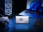 A lifestyle shot of the white SSD reader placed centrally on a blue lit desk setup flanked by a gaming PC tower a game controller and a laptop